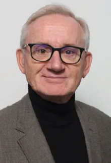 Portrait de Frédéric-Jacques Bourgade, cheveux blancs, lunettes noires, col roulé sombre et veste claire.